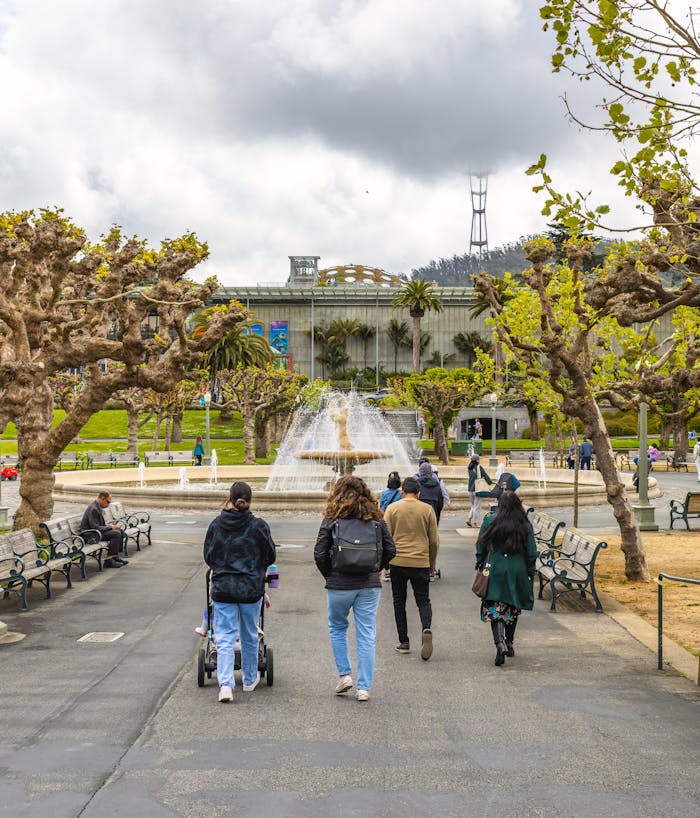 People walking towards a fountain in a picturesque urban park on a cloudy day.