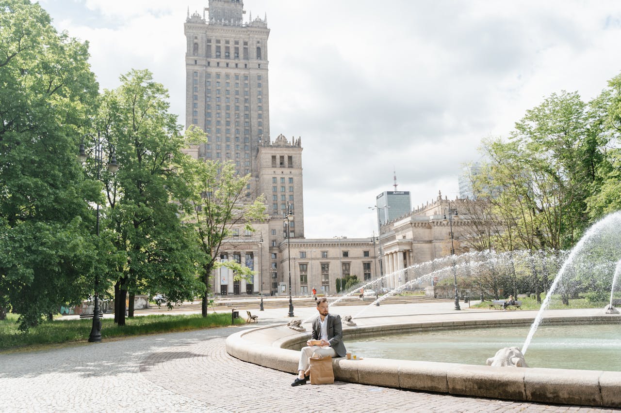 A man sits by a fountain near the Palace of Culture and Science in Warsaw, enjoying a casual lunch outdoors.